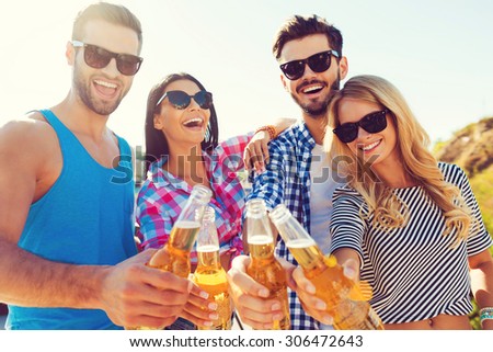 Celebrating summer. Low angle view of four cheerful young people bonding to each other and stretching out bottles with beer while standing on the roof
