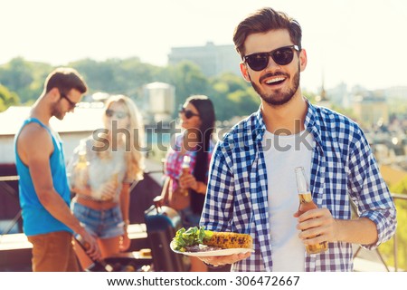 Good food with best friends. Smiling young man holding bottle with beer and plate with food while three people barbecuing in the background