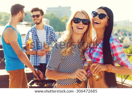 Having great time together. Two cheerful young women clinking glasses with beer and smiling while two men barbecuing in the background