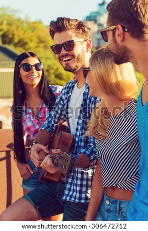 Enjoying time together. Side view of four young people bonding to each other and looking at camera while standing on the roofwith guitar