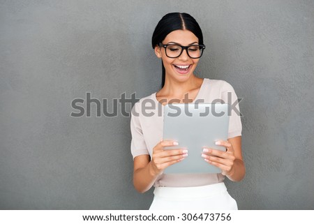 Job is done well! Joyful young businesswoman holding digital tablet and looking at it while standing against grey background