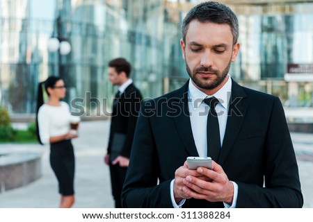 Business texting. Serious young businessman holding mobile phone and looking at it while two his colleagues talking to each other in the background