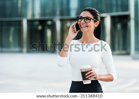 Good business talk. Cheerful young businesswoman talking on the mobile phone and holding cup of coffee while standing outdoors