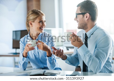 Sharing fresh news. Two joyful young people in formalwear holding cups of coffee and discussing something while working together