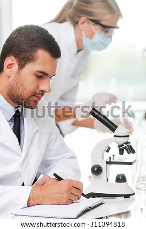 Our goal is to find the cure. Side view of male scientist writing in note pad while his female colleague making experiment in the background