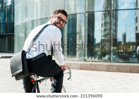 Green way to get to office. Rear view of joyful young businessman looking at camera and smiling while riding on his bicycle with office building in the background