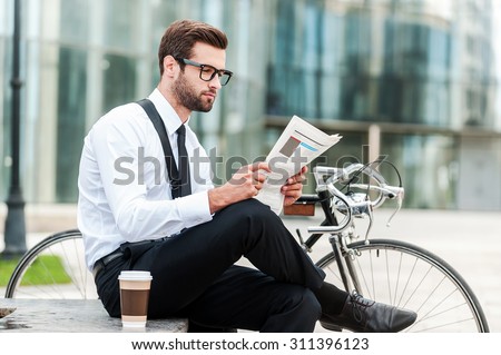 Reading the latest news. Side view of young businessman reading newspaper while sitting near his bicycle with office building in the background