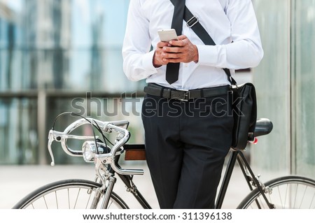 Staying in touch. Close-up of young businessman holding mobile phone while leaning at his bicycle with office building in the background