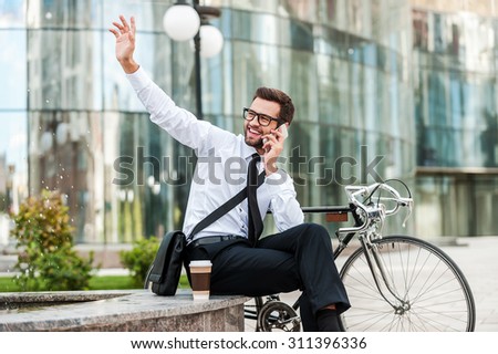 I am here! Cheerful young businessman talking on the mobile phone and waving somebody while sitting near his bicycle with office building in the background