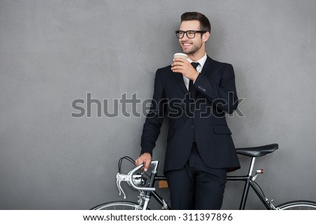 Successful businessman. Smiling young man leaning at his bicycle and holding cup of coffee while standing against grey background