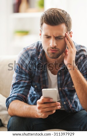 Dealing with bad news. Frustrated young man holding mobile phone and touching his forehead while sitting on sofa