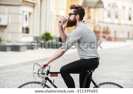 Coffee on the go. Side view of young bearded man drinking coffee while sitting on his bicycle outdoors