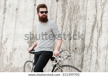 Street style. Pensive young bearded man leaning at his bicycle and looking away while standing against the concrete wall