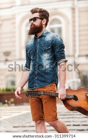 He is never far from his guitar. Low angle view of young bearded man holding guitar and looking away while walking outdoors