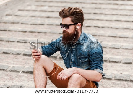 On the city wave. Top view of young bearded man in headphones holding mobile phone while sitting outdoors