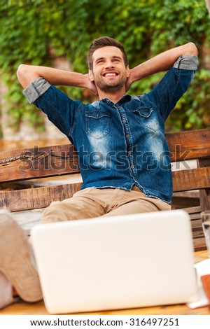 Feeling confident and relaxed. Cheerful young man holding head in hands and smiling while leaning at the wooden bench outdoors