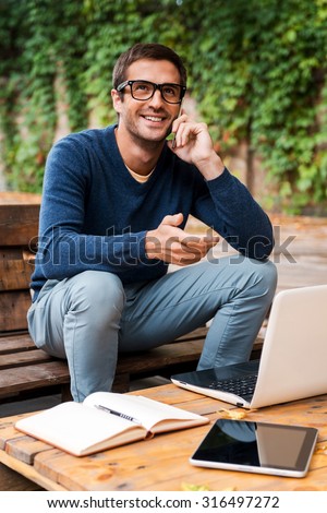 Good business talk. Happy young man talking on the mobile phonewhile sitting at the wooden table outdoors
