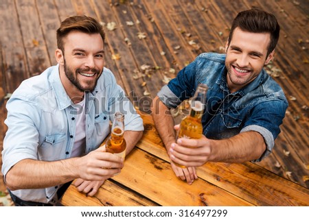 Cheers! Top view of two joyful young men stretching out bottles with beer and looking at camera while standing outdoors