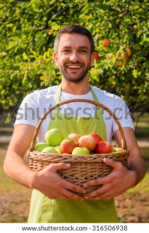 Rich harvest. Happy young gardener holding basket with apples and smiling while standing in the garden