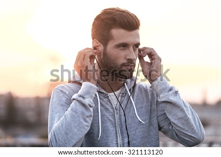Music is always with me. Confident young man putting headphones into his ears and looking away while standing outdoors