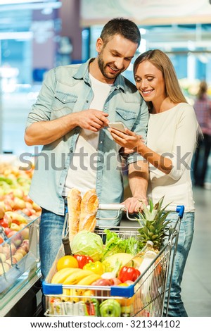 We bought all we need. Happy young couple looking at mobile phone together while standing near shopping cart in food store