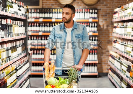 Choosing the best wine for dinner. Confident young man with shopping cart choosing wine while walking across wine store