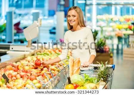 Choosing healthy food. Smiling young woman holding apple and looking at camera while standing in a food store