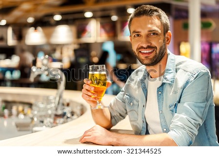 Beer time. Cheerful young man holding glass of beer and looking at camera while sitting at the bar counter