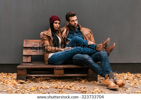 Enjoying time together. Beautiful young couple bonding to each other while sitting on the wooden pallet with grey wall in the background and fallen leaves on the floor