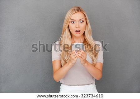 Shocking message. Surprised young blond hair woman holding mobile phone and staring at it while standing against grey background