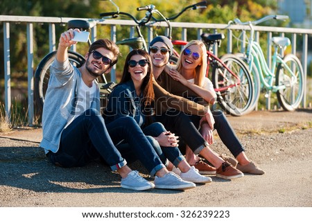 Capturing fun. Group of young smiling people bonding to each other and making selfie by smart phone while sitting outdoors together with bicycles in the background