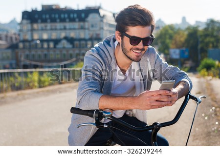 Inviting friend for a ride. Handsome young smiling man leaning at his bicycle and looking at his mobile phone while standing outdoors