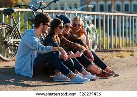 Relaxing after day riding. Group of young smiling people bonding to each other and looking at smart phone while sitting outdoors together with bicycles in the background