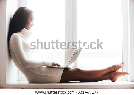 Beauty with laptop. Attractive young African woman in warm sweater working on laptop while sitting on the window sill