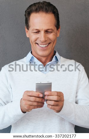 Texting to friend. Confident mature man holding mobile phone and looking at it with smile while standing against grey background