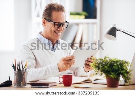 Technologies making life easier. Cheerful mature man holding digital tablet and looking at it with smile while sitting at his working place