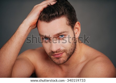 Confident and handsome. Confident young shirtless man looking at camera and holding hand in hair while holding hand on chin and sitting against grey background