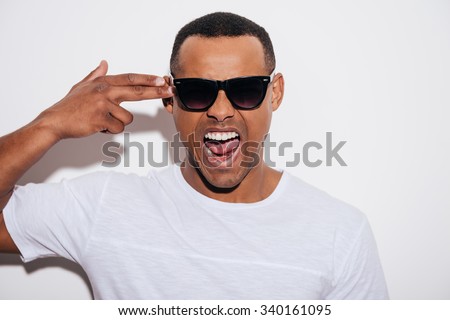 Bang! Furious young African man in sunglasses gesturing finger gun near head and keeping mouth open while standing against white background