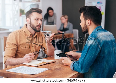 Finding solution together. Two confident young men talking and gesturing while sitting at the desk in office with two colleagues working in the background