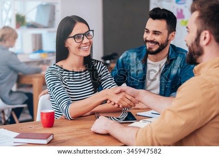 Welcome on board! Confident young woman and man shaking hands and smiling while sitting at the desk in office