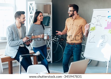 Discussing some new project together. Confident young man standing near whiteboard and pointing it with smile while his colleagues standing near him