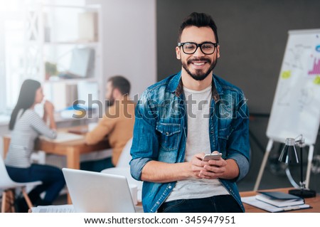 Full of new great ideas. Confident young man holding smart phone and looking at camera with smile while his colleagues working in the background