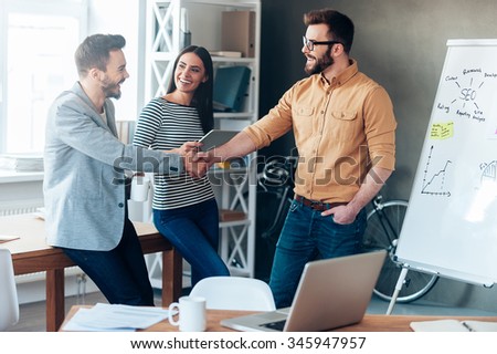 Good job! Confident young man standing near whiteboard and shaking hand to his colleague while young woman standing near them and smiling