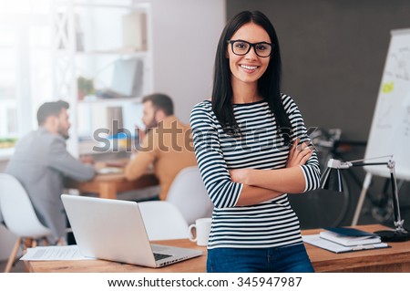 Confident and successful leader. Confident young woman keeping arms crossed and looking at camera with smile while her colleagues working in the background