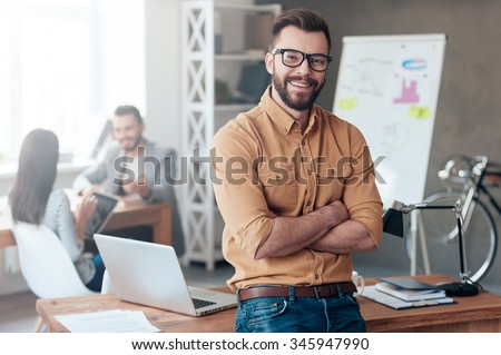 Confident team leader. Confident young man keeping arms crossed and looking at camera with smile while his colleagues working in the background