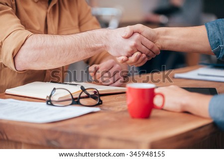 Sealing a deal. Close-up of two men shaking hands while sitting at the wooden desk