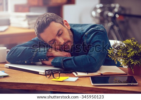 Sleeping at working place. Young man leaning his head on desk and keeping eyes closed while sitting at his working place