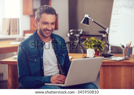 Surfing the net in office. Confident young man working on laptop and smiling while sitting at his working place in office