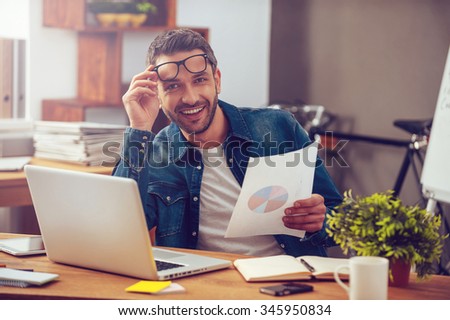 That was a great month! Handsome young man holding paper with colorful diagram and looking at camera with smile while sitting at his working place in office