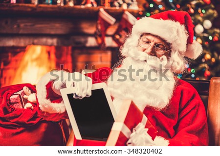 The best present! Cheerful Santa Claus putting a digital tablet into the gift box and smiling while sitting at his chair with Christmas Tree in the background